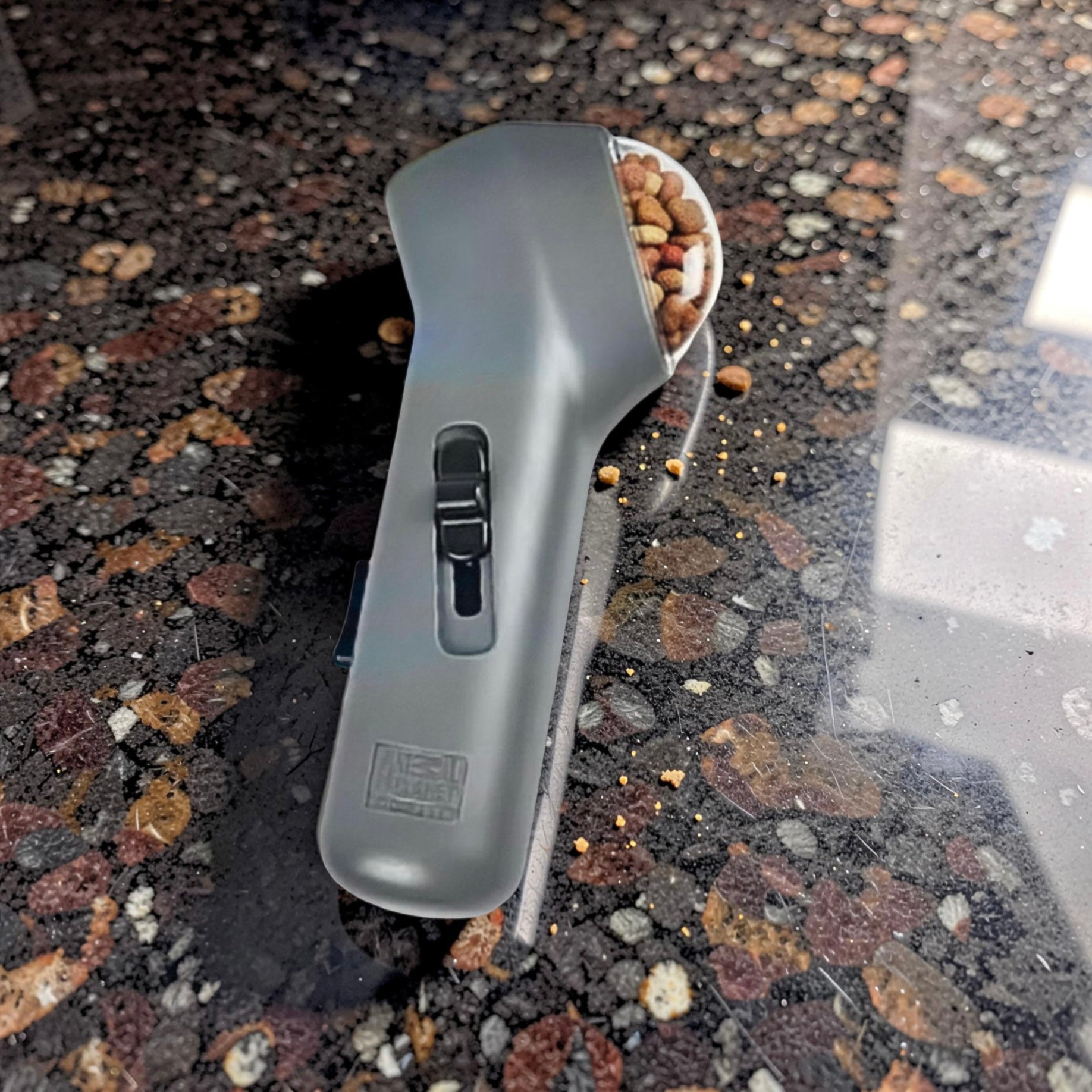 Grey pet treat launcher loaded with dog treats resting on shiny kitchen work top with crumbs around it — dog training treat shooter